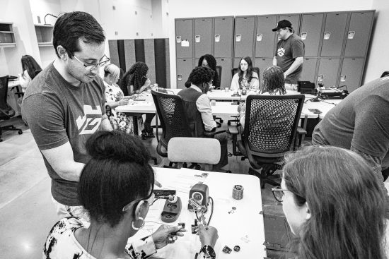 Georgia Cyber Center employee assists two young women with a soldering project in the foreground while in the background another Georgia Cyber Center employee assists a young woman with the same soldering project.