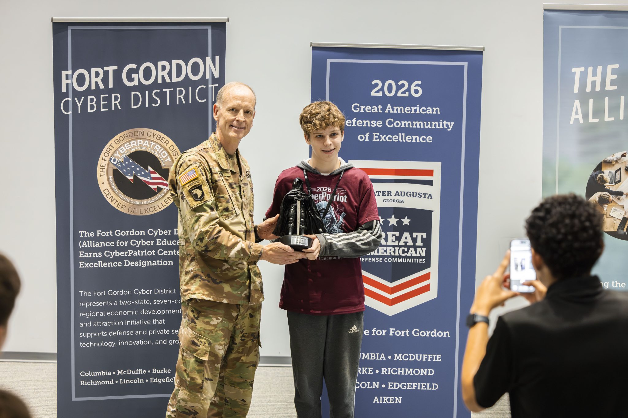 Major General Miles presenting a black trophy to a young male student wearing a maroon CyberPatriot t-shirt in front of a "Fort Gordon Cyber District" banner while someone takes a photo in the foreground.