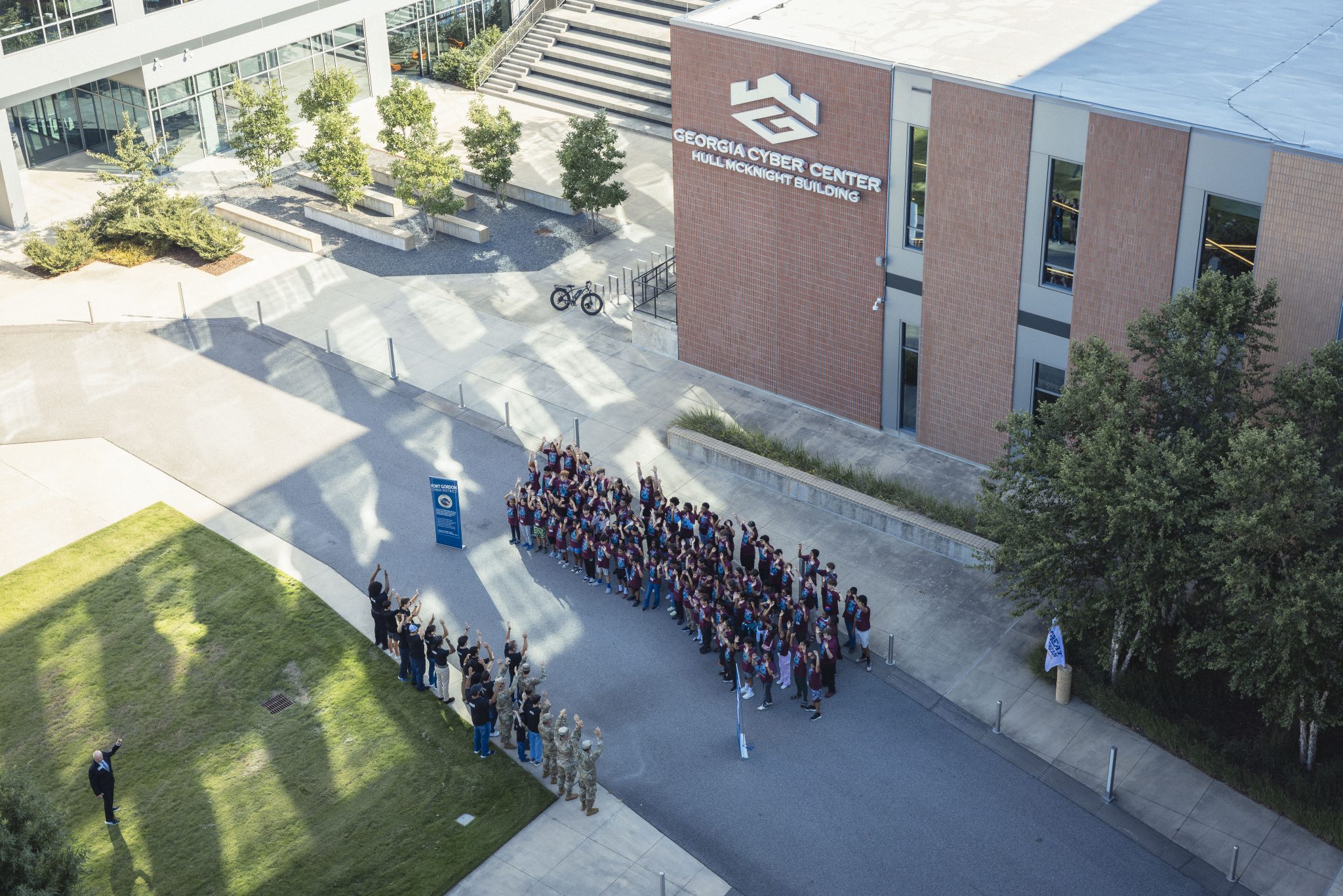 High-angle aerial shot of a large group of students in matching t-shirts lined up on the pavement outside the brick Georgia Cyber Center Hull McKnight Building.