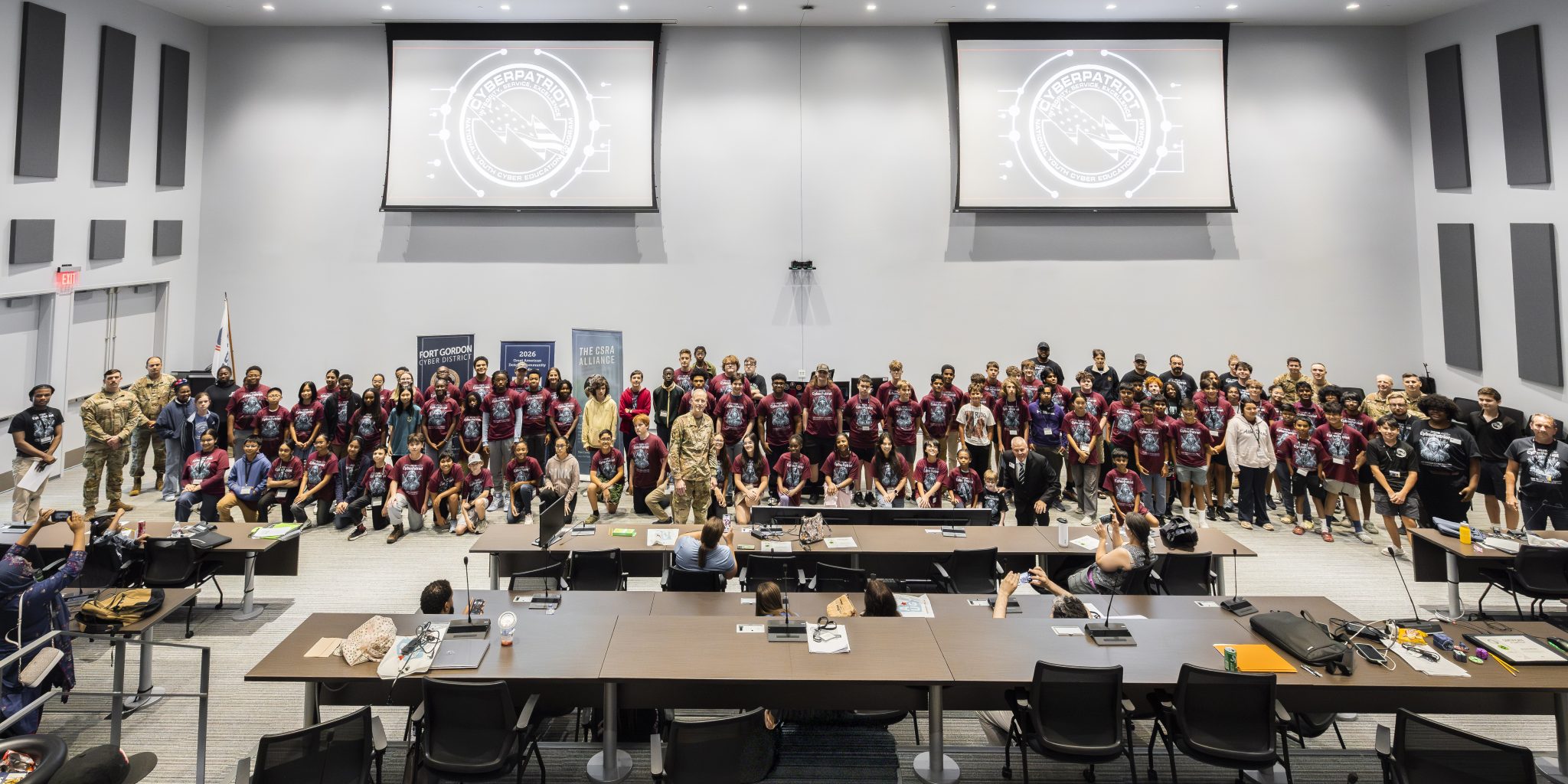 CyberPatriot 2026 Advanced campers stand at the front of the Plug n Play auditorium at the Georgia Cyber Center alongside military officials for graduation ceremony