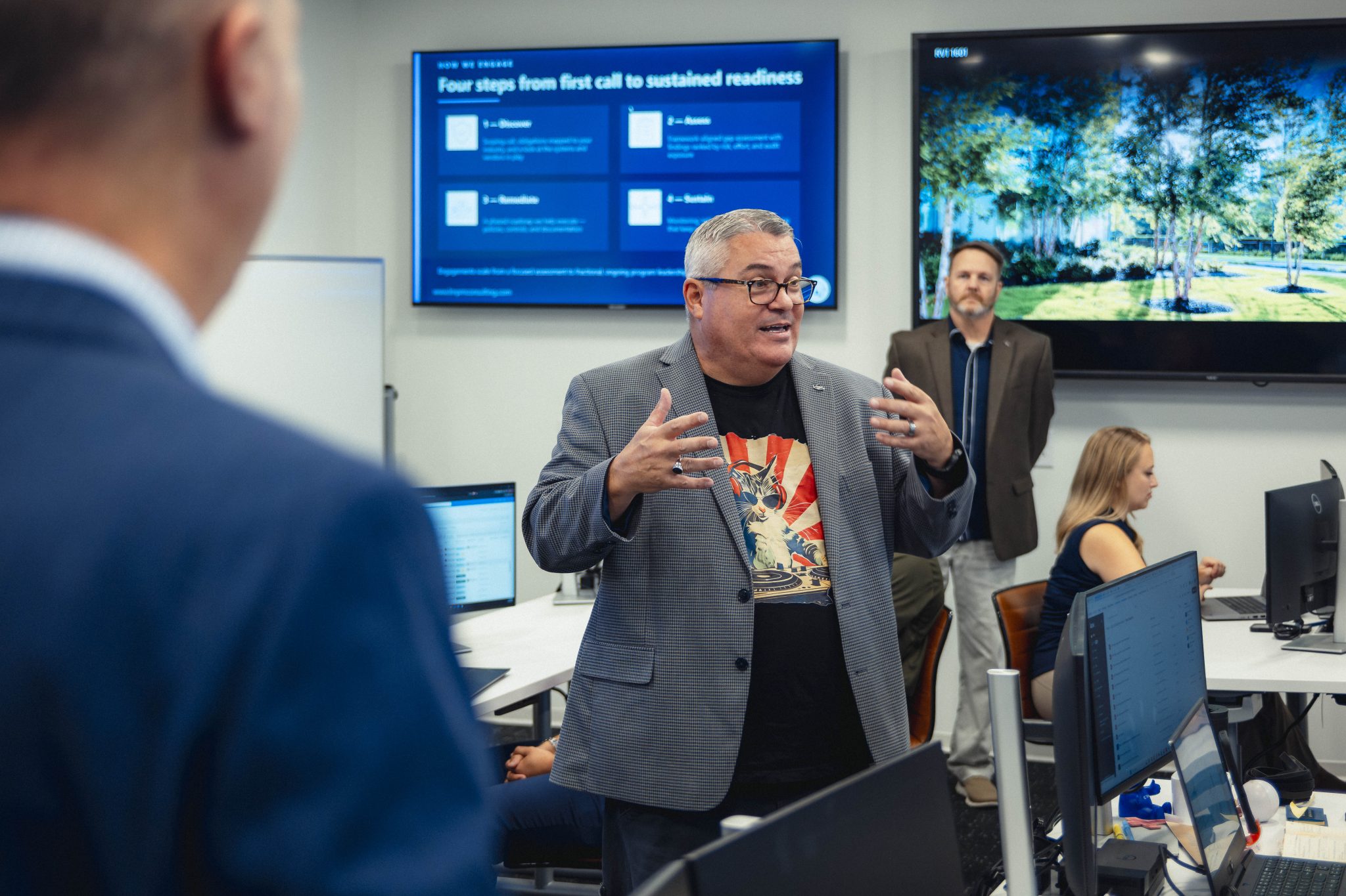 John McLain speaks with hands outstretched inside a modern tech room while wearing glasses, a grey blazer, and a cat graphic t-shirt.