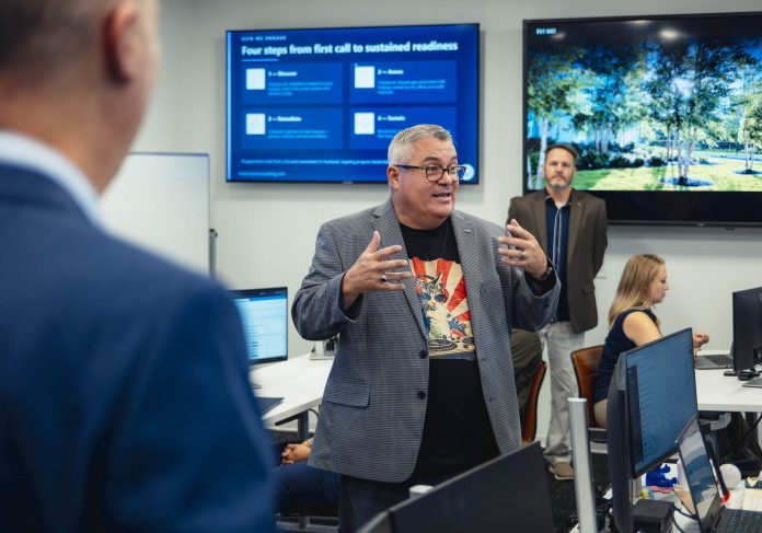 John McLain speaks with hands outstretched inside a modern tech room while wearing glasses, a grey blazer, and a cat graphic t-shirt.