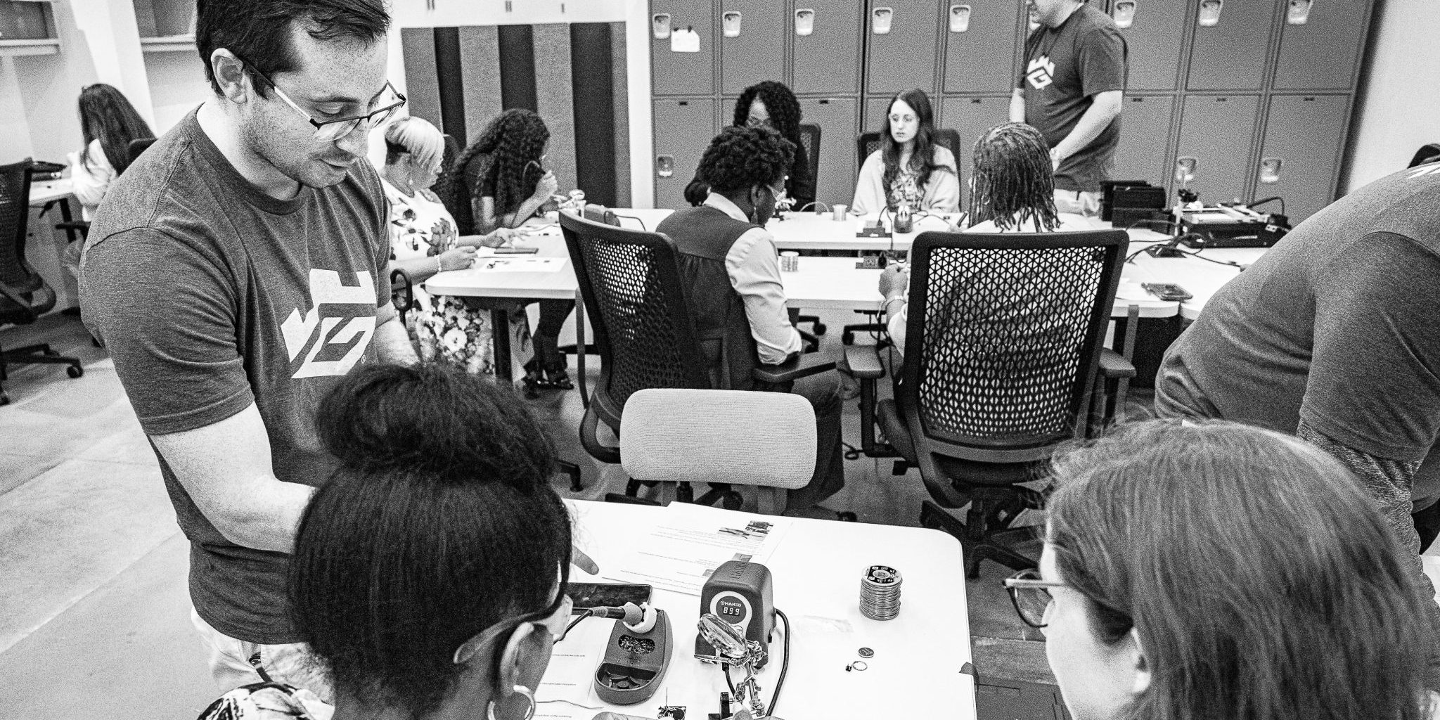 Georgia Cyber Center employee assists two young women with a soldering project in the foreground while in the background another Georgia Cyber Center employee assists a young woman with the same soldering project.